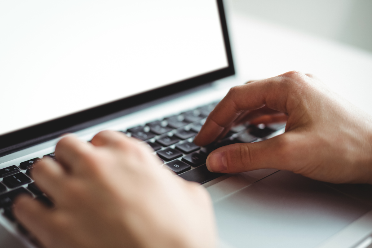 Mãos posicionadas sobre teclado preto de notebook prateado com tela em branco iluminada ao fundo.