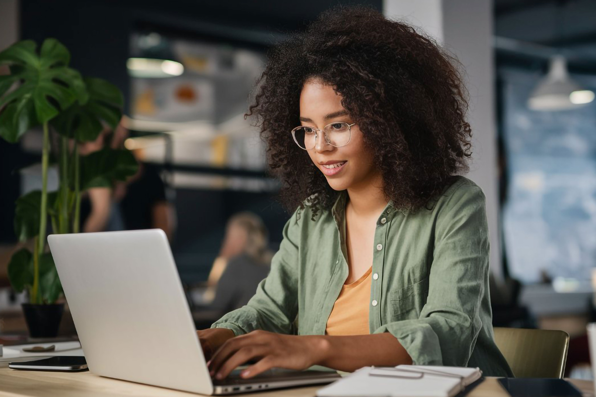 Mulher jovem de cabelos cacheados e óculos digita em laptop cinza em ambiente corporativo com plantas