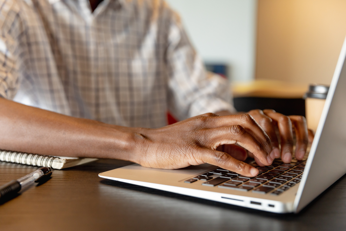 Mãos de pessoa negra digitando em teclado de laptop prateado, com caderno espiral e caneta ao lado sobre mesa escura