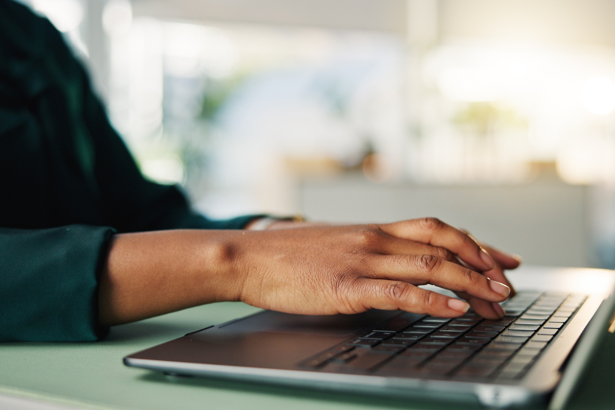 Mão de mulher negra digitando em teclado de notebook cinza sobre mesa verde com luz natural ao fundo