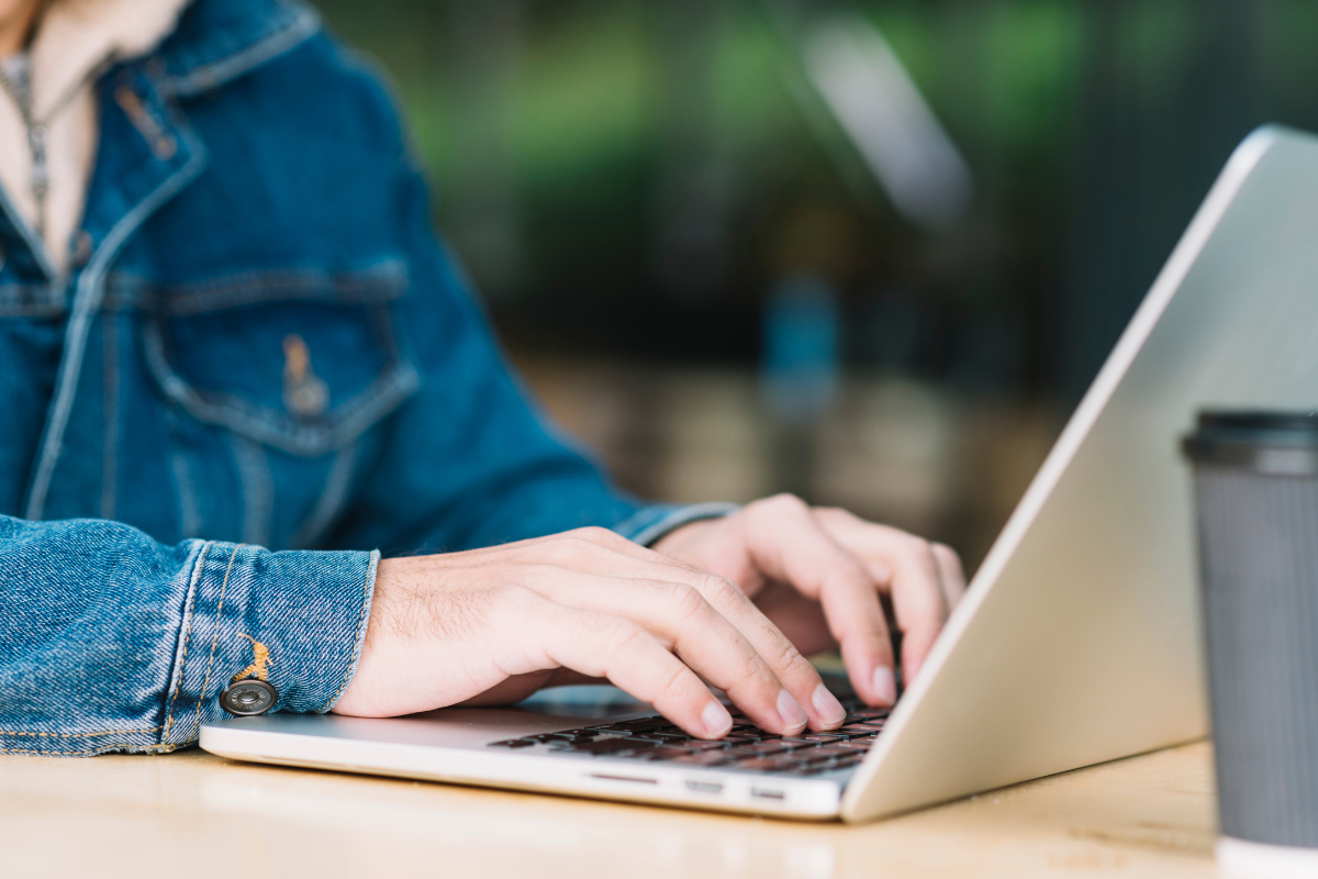 Mãos digitando em laptop prateado sobre mesa de madeira; pessoa usa jaqueta jeans azul, copo cinza ao lado.