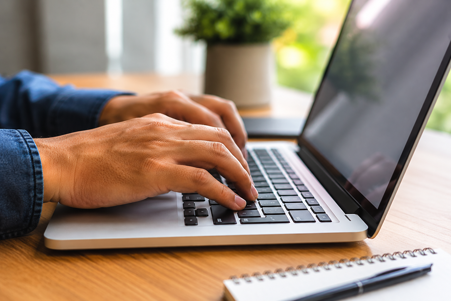 Mãos masculinas com camisa jeans digitando em teclado de laptop prateado sobre mesa de madeira com caderno e planta.