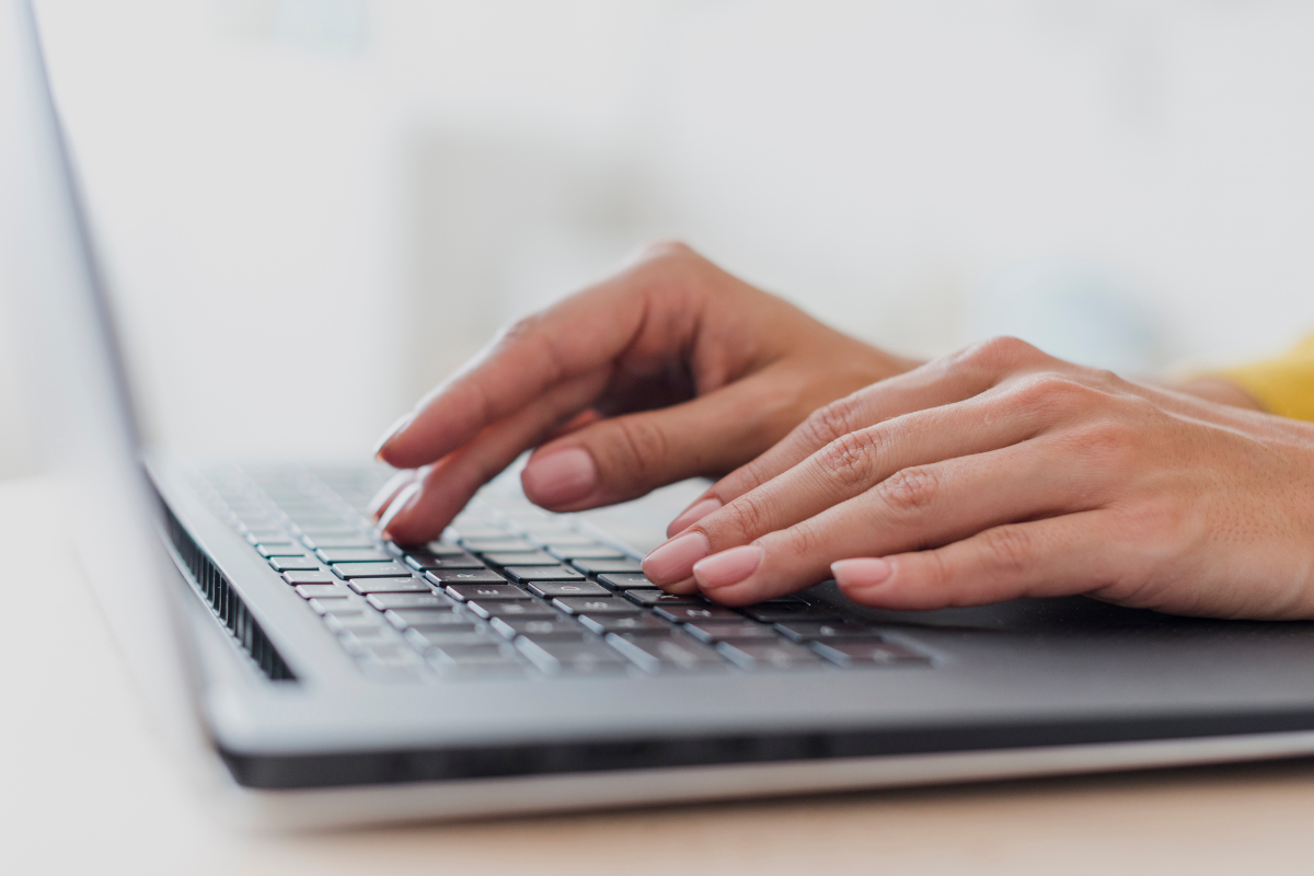 Mãos femininas com unhas naturais digitando em teclado de notebook cinza sobre mesa branca.