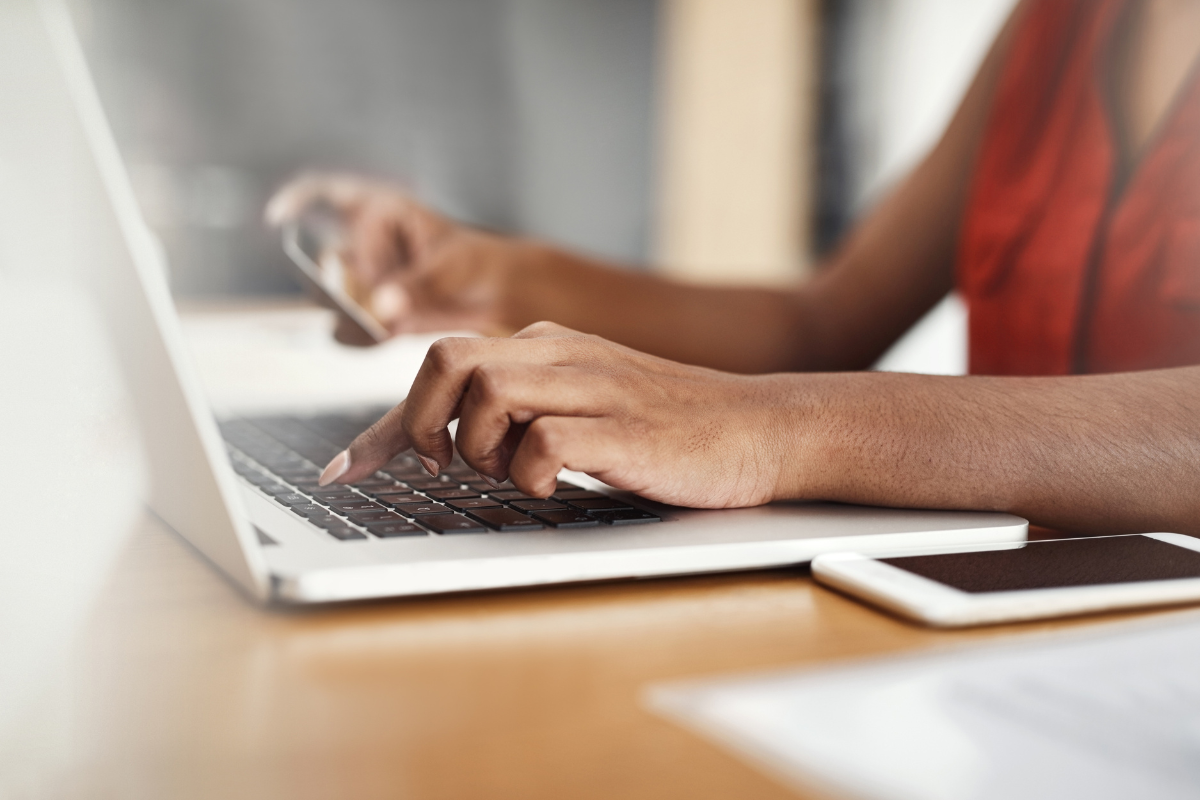 Mãos femininas digitando em laptop prateado enquanto segura celular, mesa de madeira com tablet ao lado