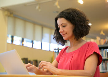 Mulher de blusa vermelha digita em notebook prateado sentada em ambiente de biblioteca com estantes de livros ao fundo