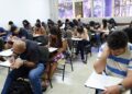 Sala de aula com diversos candidatos sentados em carteiras realizando prova em papel