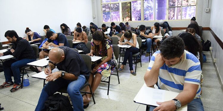 Sala de aula com diversos candidatos sentados em carteiras realizando prova em papel