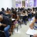 Sala de aula com diversos candidatos sentados em carteiras realizando prova em papel