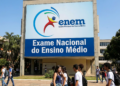 Jovens estudantes circulam em frente a painel do ENEM com logo e texto "Exame Nacional do Ensino Médio".