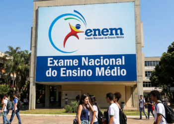 Jovens estudantes circulam em frente a painel do ENEM com logo e texto "Exame Nacional do Ensino Médio".