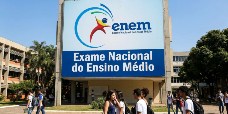 Jovens estudantes circulam em frente a painel do ENEM com logo e texto "Exame Nacional do Ensino Médio".