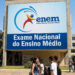 Jovens estudantes circulam em frente a painel do ENEM com logo e texto "Exame Nacional do Ensino Médio".