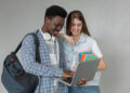 Dois estudantes sorriem ao consultar um notebook enquanto a jovem segura cadernos coloridos em fundo neutro