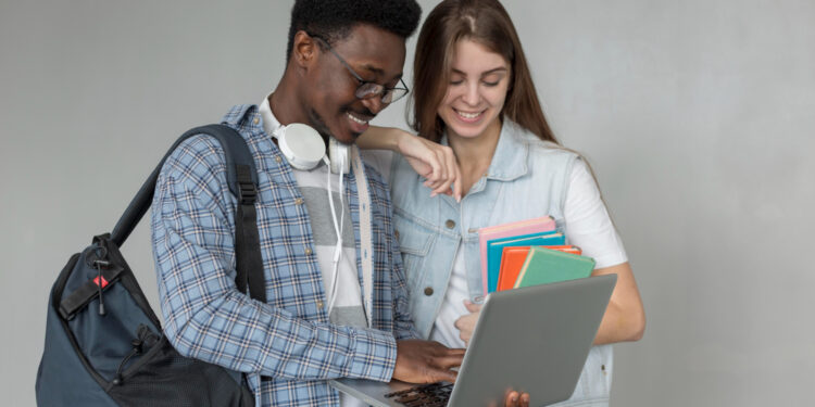 Dois estudantes sorriem ao consultar um notebook enquanto a jovem segura cadernos coloridos em fundo neutro