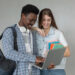 Dois estudantes sorriem ao consultar um notebook enquanto a jovem segura cadernos coloridos em fundo neutro