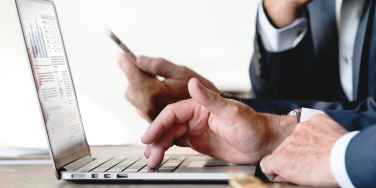 Empresário utilizando notebook e celular para consultas digitais em ambiente corporativo