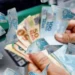 Cédulas de R$ 100 sobre imagem de dinheiro em mãos, ilustrando pagamento do INSS acima de R$ 8 mil do INSS