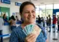 Mulher sorrindo segurando cédulas de real em agência da Caixa, representando a liberação de R$ 1.078,01.