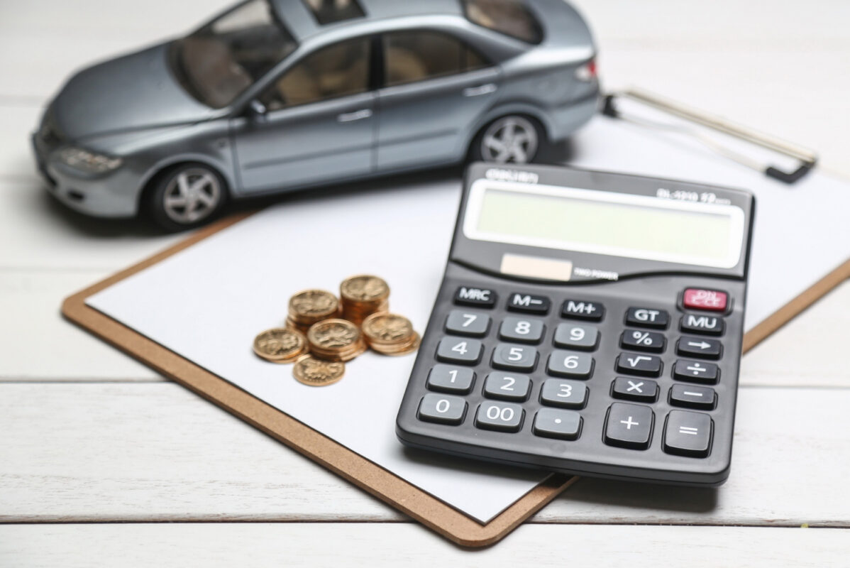Calculadora preta ao lado de moedas douradas e miniatura de carro cinza sobre prancheta de madeira