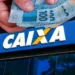 Mão masculina segurando cédulas de R$ 100 reais na cor azul diante de letreiro da Caixa Econômica Federal