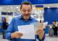 Homem sorrindo, segurando dinheiro e documento em agência bancária, representando pagamento da restituição do Imposto de Renda nesta sexta-feira (29).