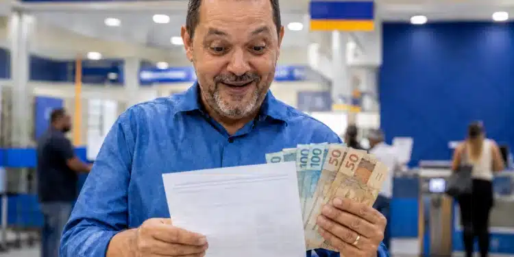 Homem sorrindo, segurando dinheiro e documento em agência bancária, representando pagamento da restituição do Imposto de Renda nesta sexta-feira (29).