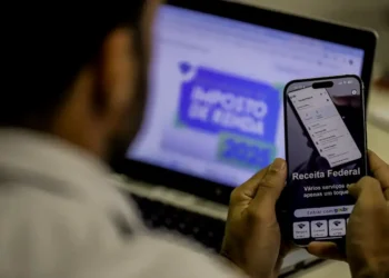 Pessoa acessando serviços da Receita Federal pelo celular enquanto declara o imposto de renda 2026 no computador.