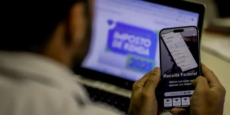 Pessoa acessando serviços da Receita Federal pelo celular enquanto declara o imposto de renda 2026 no computador.