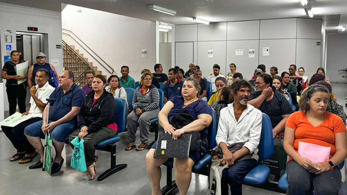 Grupo de pessoas aguarda atendimento sentado em sala de espera de agência do INSS, com documentos nas mãos.