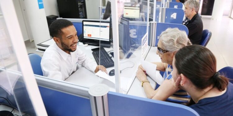 Atendente sorri ao atender duas pessoas em guichê de agência do INSS.