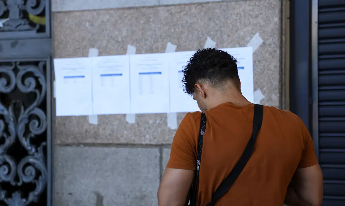 Jovem de camiseta laranja e alça preta confere listas de papéis coladas em mural de pedra, de costas para a câmera