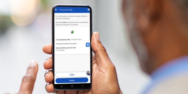 Homem idoso segura celular com a tela do serviço Prova de Vida do aplicativo Meu INSS aberta exibindo dados do benefício