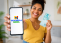 Mulher sorridente exibe smartphone com logo Desenrola Brasil e segura nota de R$ 100 em ambiente doméstico.