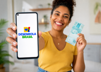 Mulher sorridente exibe smartphone com logo Desenrola Brasil e segura nota de R$ 100 em ambiente doméstico.