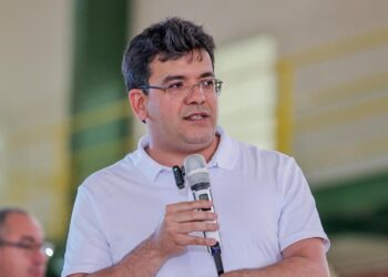 Rafael Fonteles, governador do Piauí desde 2023 , de camisa polo branca e óculos, segura microfone durante discurso em ginásio