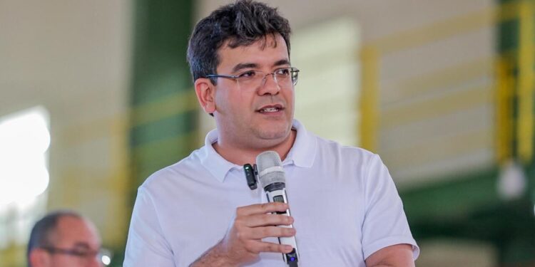 Rafael Fonteles, governador do Piauí desde 2023 , de camisa polo branca e óculos, segura microfone durante discurso em ginásio