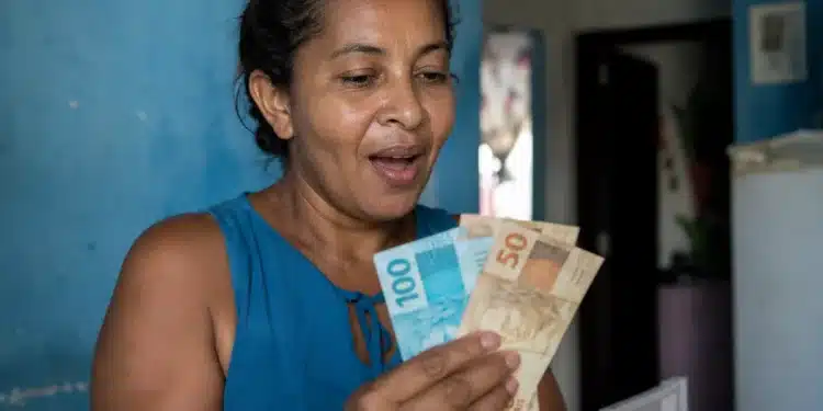 Mulher segurando cédulas de dinheiro e conferindo valor, representando o novo lote do auxílio de R$ 7,3 mil liberado pelo governo.