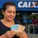 Mulher brasileira segurando notas de reais em frente à Caixa, representando o pagamento de R$ 2.100 em 2026.