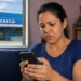 Mulher preocupada olhando o celular dentro de casa, com agência da CAIXA ao fundo, representando alerta sobre procedimento obrigatório para receber R$ 300 em junho.