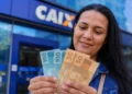 Pessoa segurando notas de R$ 100 e R$ 50 em frente a uma agência da Caixa, representando o pagamento de R$ 1.128,01 em maio.
