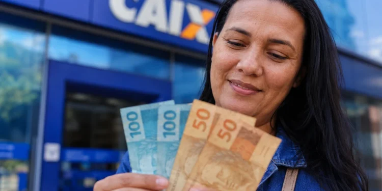 Pessoa segurando notas de R$ 100 e R$ 50 em frente a uma agência da Caixa, representando o pagamento de R$ 1.128,01 em maio.