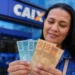 Pessoa segurando notas de R$ 100 e R$ 50 em frente a uma agência da Caixa, representando o pagamento de R$ 1.128,01 em maio.