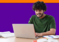 Jovem de camiseta verde sorri enquanto usa notebook sobre mesa com livros e cadernos de estudo.