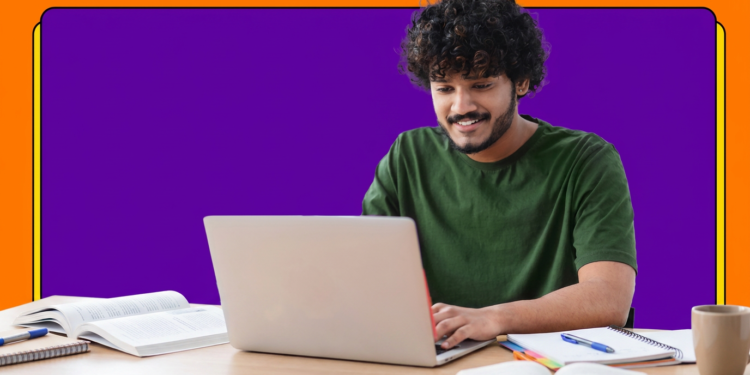 Jovem de camiseta verde sorri enquanto usa notebook sobre mesa com livros e cadernos de estudo.