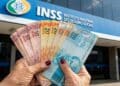 Mãos de beneficiária do INSS seguram cédulas em leque em frente à fachada de uma agência da Previdência Social