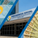 Montagem em três faixas diagonais com cédulas de cinquenta e cem reais, o logotipo da Previdência e a fachada do edifício-sede do INSS