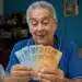 Idoso aposentado segurando notas de reais dentro de casa representando a confirmação de pagamentos de R$ 8.475,55 do INSS e segunda parcela do 13º salário.