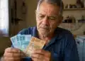 Aposentado segurando uma nota de 100 e outra de 50 reais, com expressão preocupada, representando os pagamentos menores do INSS em junho.
