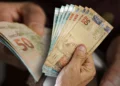 Pessoa segurando notas de R$ 50 em dinheiro, representando o pagamento de R$ 1.081 que será liberado nesta semana para CPFs com finais de 0 a 9.