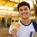 Estudante segurando notas de reais em frente a uma escola pública, representando o pagamento do programa Pé-de-Meia nesta terça-feira (26).